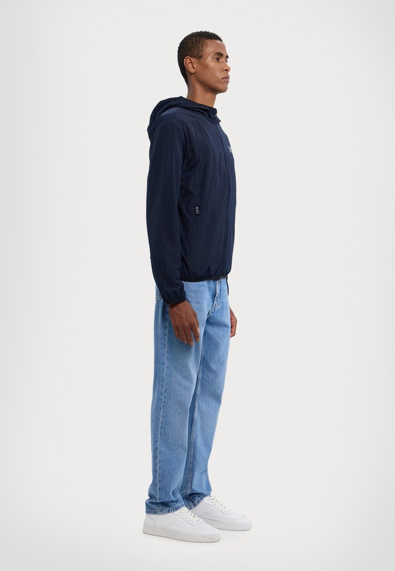 Marineblå hættejakke med en lynlåslomme, elastiske manchetter og et let design, parret med lyseblå jeans og hvide sneakers.