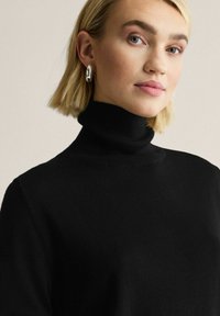 Nainen, jolla on lyhyet vaaleat hiukset, hopearenkaat korvissa ja musta pooloneule, katsoo hieman oikealle, neutraali tausta.