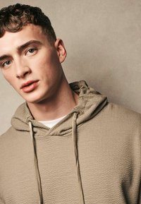 Khaki-farbenes Sweatshirt mit strukturierter Oberfläche und Kapuze mit Kordelzug, das ein horizontales Muster aufweist und ein weißes Hemd darunter trägt. Nahaufnahme.