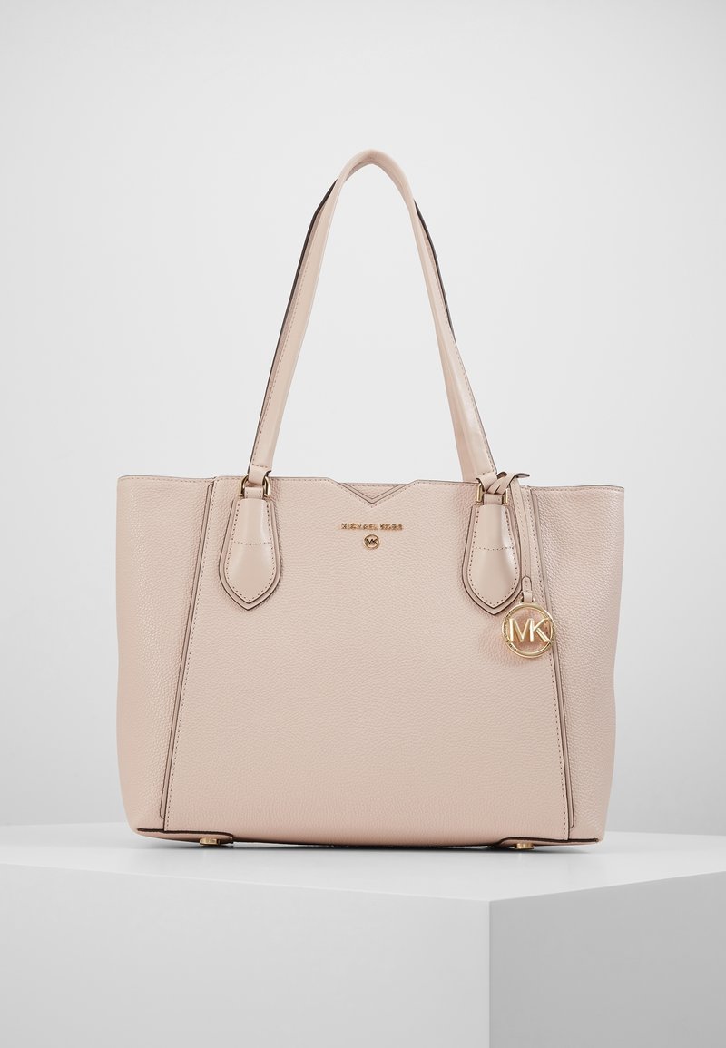 Sac en cuir texturé rose clair avec doubles anses, plaque en or avec le logo Michael Kors et charme MK circulaire suspendu sur un piédestal blanc.