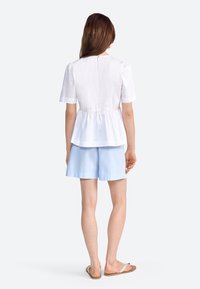 Hvid kortærmet bluse med peplum-kant, parret med lyseblå shorts. Blusen har en rund halsudskæring og en knaplukning på ryggen.