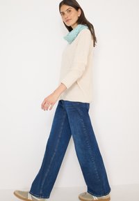 Beige gebreide trui, blauwe wijde denimbroek en een lichtblauwe patroon sjaal. De outfit heeft relaxte vormen en contrasterende texturen.