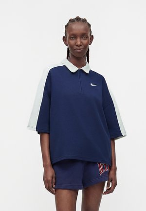 Femme aux cheveux tressés portant un polo bleu marine à manches courtes avec des manches blanches et un short bleu marine assorti avec un texte rouge.