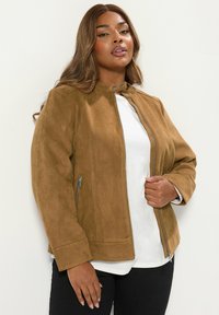 Brun mockajacka med ståkrage, dragkedjor, sidofickor och en slät yta, bärs över en vit skjorta och svarta byxor.