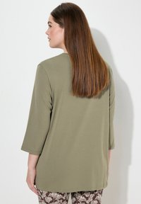 Olijfgroene, langemouwenshirt met een ontspannen pasvorm en een afgeronde zoom. Glad textuur, geen zichtbare patronen. Gedragen met een geblokte bruine broek.