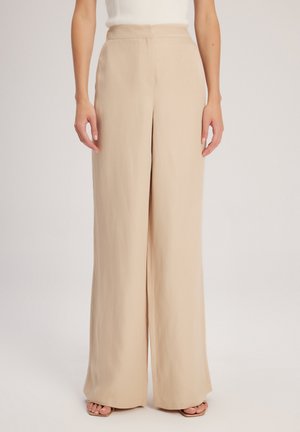 Femme portant un pantalon beige à taille haute et jambes larges, associé à un haut blanc sans manches, debout devant un fond clair uni.