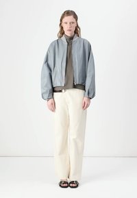 Casaco bomber cinza claro com uma textura suave, apresentando um fecho éclair à frente e bolsos laterais, combinado com uma parte de cima escura e calças largas cor creme.