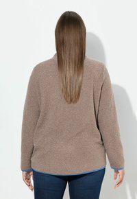 Brauner, flauschiger Pullover mit langen Ärmeln und abgerundetem Saum, mit blauen Akzenten an den Bündchen, über einem blauen Hemd geschichtet, von hinten betrachtet.