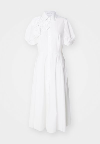Vit skjortklänning med korta puffärmar, med blommig applikation på vänster axel och en flödande, ankellång kjol.