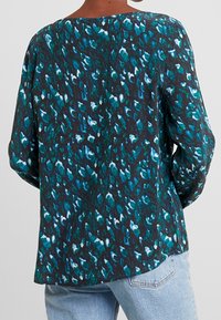 Kobieta w luźnej bluzce z długim rękawem w abstrakcyjny wzór w odcieniach turkusu, czerni i bieli, zestawiona z jasnoniebieskimi dżinsami, pokazana od tyłu.