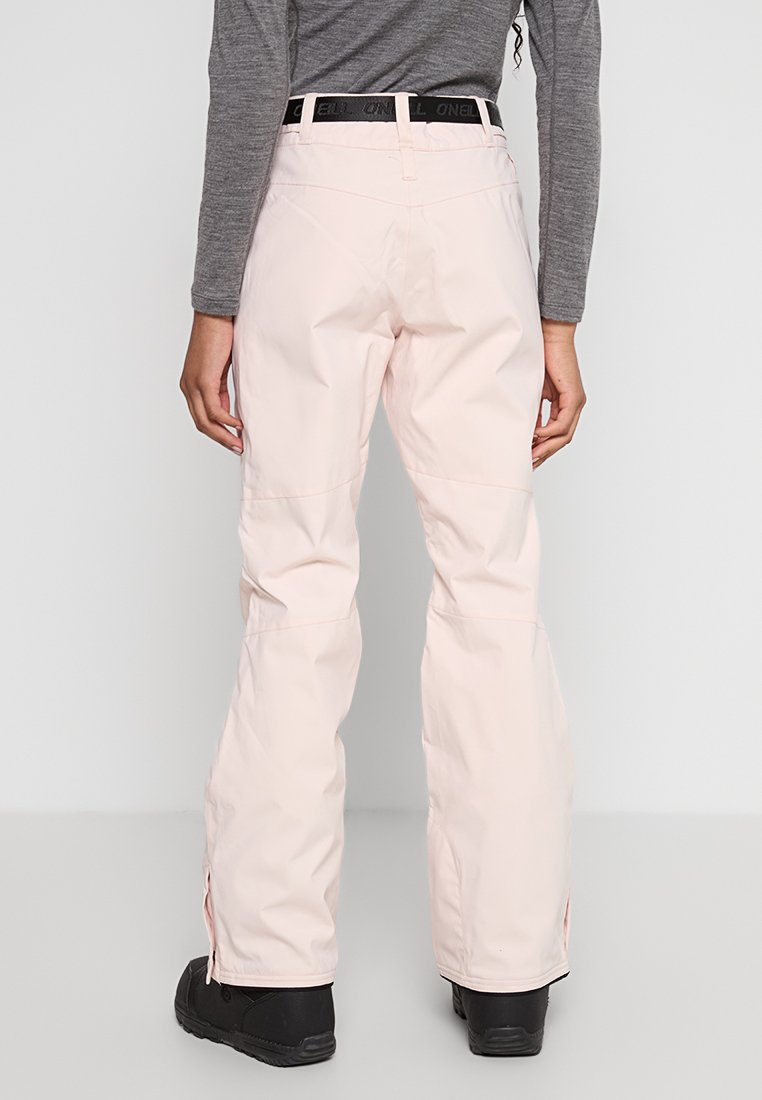 Osoba w jasnoróżowych spodniach narciarskich, z czarnym paskiem, szarym topem z długim rękawem i czarnymi butami stoi na jednolitym tle.