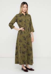 Giovane donna in un vestito lungo olive green con fiori, a bottoni e maniche a tre quarti, con ballerine rosso scuro, in posa su uno sfondo semplice.
