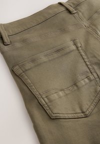 Detail der Gesäßtasche an olivgrünen Hosen, die Nähte, Gürtelschlaufen und Stoffstruktur in Nahaufnahme zeigt.
