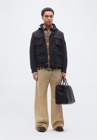 Άνδρας που φοράει μαύρο σακάκι πάνω από καφέ πουκάμισο καρό, μπεζ παντελόνια φαρδιάς γραμμής, μαύρα παπούτσια, κρατώντας μαύρη τσάντα duffel, στέκεται σε λευκό φόντο.