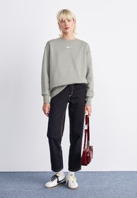 Sweatshirt cinza com decote redondo e logotipo branco, jeans pretos de perna reta com costuras visíveis, sapatos brancos com detalhes em preto e amarelo, bolsa de ombro vermelha.