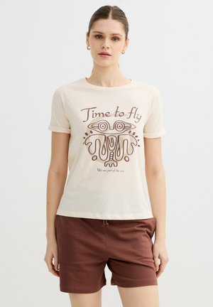 Joven mujer con el pelo recogido, vistiendo una camiseta color crema con la frase "Time to fly" y un diseño abstracto de un pájaro, combinada con pantalones cortos marrones sobre un fondo blanco.