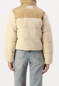 Kurtka puchowa z kremowym zewnętrzem ze skóry baraniej oraz górną częścią z nylonu w kolorze beżowym. Posiada kaptur i elastyczne mankiety, prezentując krótki fason.