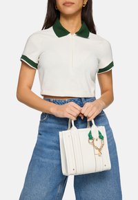 Vit mini-handväska med texturerat material, dubbla handtag upptill, grön dekorativ hårdvara och ett guldkedjeaccent. Bärs med en kort poloöverdels.