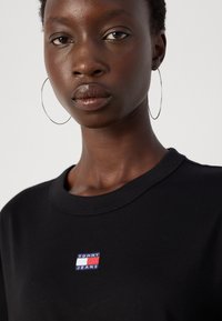 Tricou din bumbac negru, cu un mic logo brodat "TOMMY JEANS" în culorile albastru, roșu și alb. Design cu guler rotund și o textură netedă.