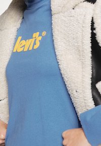 Světle modrá košile s dlouhými rukávy a žlutým logem "Levi's", pod kterou je navlečená krémová chlupatá bunda s černými koženými detaily.