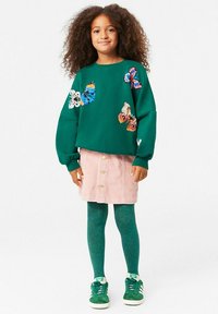 Felpa verde con applicazioni a forma di farfalla, gonna in velluto corduroy rosa con bottoni, collant teal e sneaker verdi con accenti bianchi.