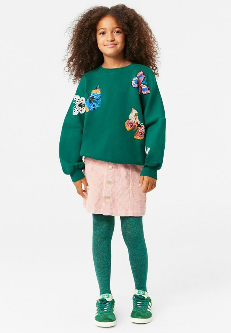 Felpa verde con applicazioni a forma di farfalla, gonna in velluto corduroy rosa con bottoni, collant teal e sneaker verdi con accenti bianchi.