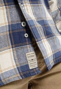 Niebieska i kremowa koszula flanelowa w kratę z zapięciem na guziki, białymi guzikami oraz naszywką z informacjami o marce. Miękka w dotyku.