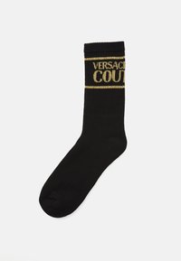 Čierna ponožka po členky s zlatým pleteným nápisom "Versace Couture" blízko vrchu, rebrovaným lemom a hladkou časťou chodidla na bielom pozadí.