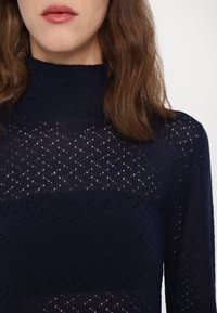 Maglia a maniche lunghe color blu navy con collo alto. Presenta un tessuto leggero e lavorato a maglia con piccoli motivi a diamante su tutta la superficie.
