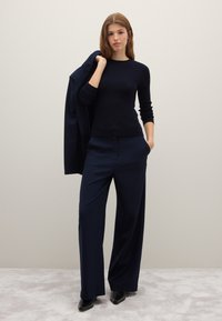 Combinaison de pull côtelé bleu marine et pantalon à jambes larges. Veste ajustée posée sur l'épaule. Texture lisse, design épuré, et schéma monochrome.