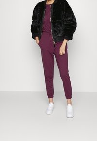 Giacca bomber in ecopelliccia nera sopra una tuta bordeaux, con pantaloni a sigaretta e polsini elastici, abbinata a scarpe da ginnastica bianche.