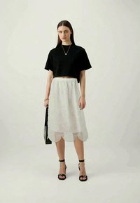 Camiseta preta corta, saia midi branca em renda com bainha recortada, sandálias pretas de tiras e mala de ombro, em pé sobre um fundo claro.