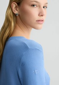 Helles blaues Strickoberteil mit strukturierten Details an den Schultern, kombiniert mit herzförmigen silbernen Ohrringen. Glattes Material mit figurbetontem Design.