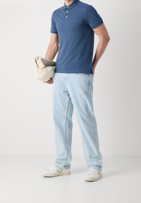 Homme portant un polo bleu, un jean bleu clair, des baskets blanches, tenant un manteau beige plié sur un fond blanc uni.