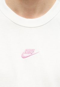 Κοντινό πλάνο σε λευκό γιακά πουκάμισου με μικρό ροζ λογότυπο Nike κεντημένο κάτω από τον γιακά.