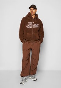 Brown fleece zip-up hoodie with a graphic logo, paired with loose brown cargo pants and gray sports shoes. Includes a matching brown cap.