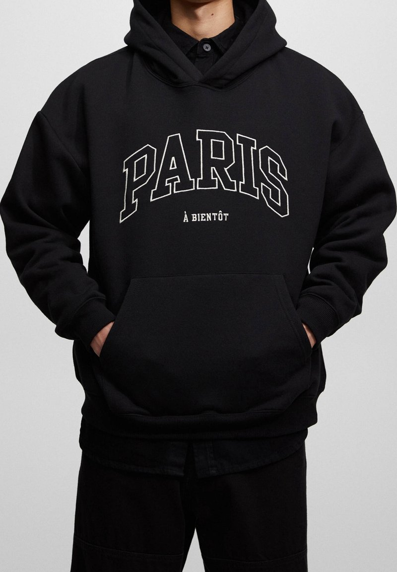 Sudadera con capucha negra y bolsillo delantero, presenta el texto blanco "PARIS" y "À BIENTÔT" en una fuente gruesa. Tela suave y texturizada.