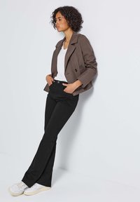 Veste brune courte, t-shirt blanc, pantalon noir évasé et baskets blanches ; tissu lisse, design simple, porté sur un fond uni.