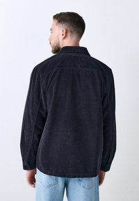 Dunkelblaue Cordjacke mit Kragen, vertikal gerippter Textur und Knöpfen an den Ärmelabschlüssen. Getragen mit hellblauen Jeans, von hinten gesehen.