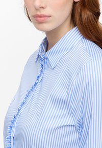 Light blue and white striped blouse with a ruffled front, button closures, and a collar. Made of smooth fabric, fitted shape.