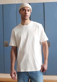 Vit bomull t-shirt med korta ärmar, lös passform, med en subtil präglad design från 1955 på framsidan. Bärs tillsammans med ljusblå jeans.