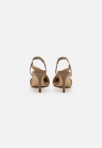 Sandales à talons beiges avec un design à lanières, présentant un talon fin et un dos arrondi. Fabriquées en matériau lisse avec des accents métalliques.