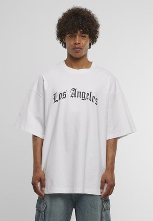 Persoană tânără cu păr creț purtând un tricou alb supradimensionat cu inscripția „Los Angeles” în font gotic negru și blugi cargo bleu deschis.