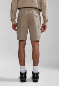 Pantalones cortos de cargo beige con bolsillos traseros, frente plano y dobladillos enrollados, combinados con zapatos negros y calcetines blancos. Tejido suave y diseño funcional.