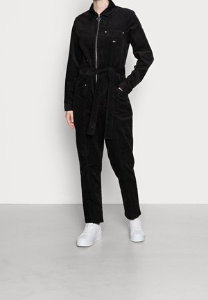 Musta corduroy-haalaripuku, jossa on pitkät hihat, kauluspääntie ja edessä vetoketju. Siinä on vyö vyötäröllä ja kaksi etutaskua. Jalkineina valkoiset tennarit.