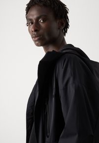 Black waterproof jacket with a zippered pocket, high collar, and textured inner lining. The material has a smooth finish.