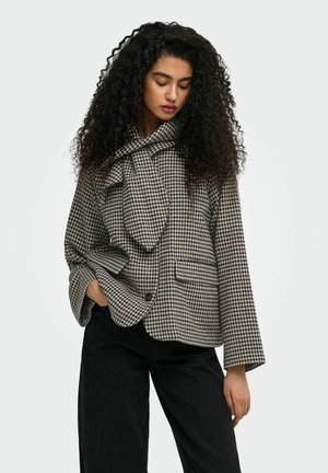 Femme aux cheveux bouclés portant une veste à motif pied-de-poule noir et blanc et un pantalon noir, une main dans sa poche.