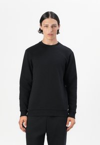 Giovane con capelli corti e scuri, che indossa una semplice felpa nera a girocollo e pantaloni neri abbinati, contro uno sfondo bianco.