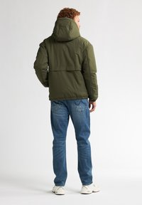 Olivgrüner, gepolsterter Jacke mit Kapuze, die einen lässigen Schnitt und gestickte Details aufweist, kombiniert mit hellblauen Jeans und weißen Sneakers.
