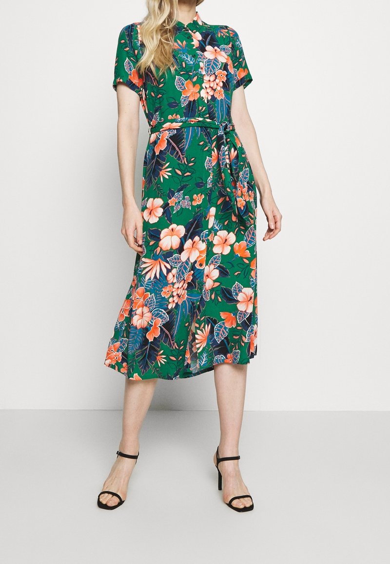 Kwiecista sukienka midi z zielonej tkaniny, krótki rękaw, pas z paskiem, w pomarańczowe i niebieskie wzory kwiatowe, z luźnym dołem i sandałami.